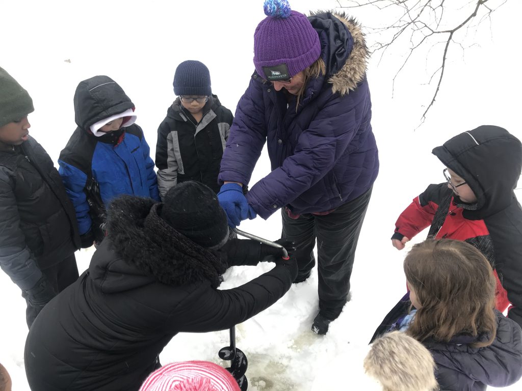 Dodge Nature Center Volunteer Leslie Krona helps elementary students drill a hole in the Farm Pond ice.