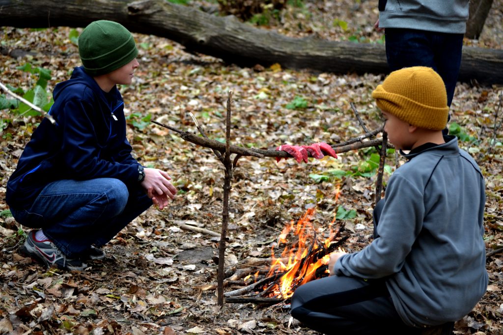 Two Dodge Nature Center campers roast strips of meat over a campfire in the woods.