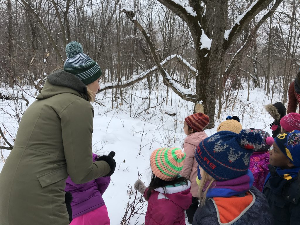 Dodge Nature Center Naturalist Fellow Julia Pedersen and her students gather in the snowy woods to observe tracks in the snow.